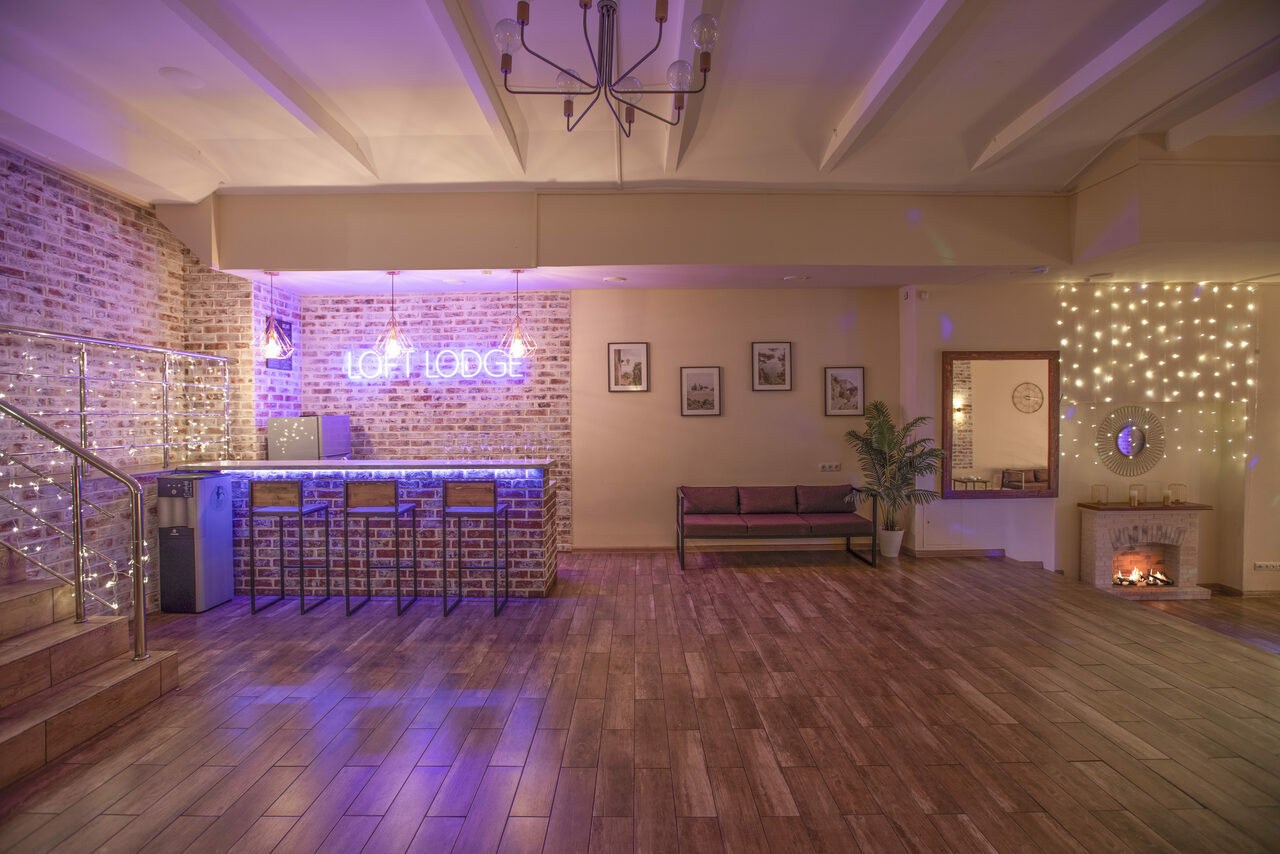Арендовать Loft Lodge (Лофт Лодж) 7. Просторный уютный лофт. Лофт-пространство с барной зоной и кирпичными стенами. На стойке — три стула, неоновая надпись "LOFT LODGE". Деревянный пол, мягкий диван, декоративный камин, картины на стене. Светодиодные гирлянды и потолочный светильник.