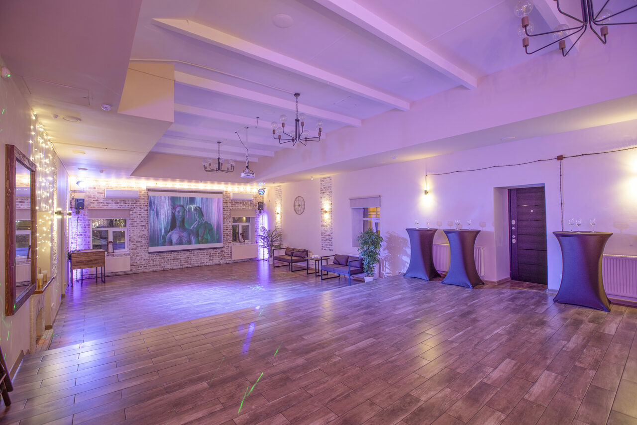Арендовать Loft Lodge (Лофт Лодж) 7. Просторный уютный лофт. Просторное помещение с деревянным полом и белыми стенами. Подвесные светильники, декорировано гирляндами. Есть несколько столиков и кресел у стены, высокий стол для ресепшн. На стене картина, рядом дверь. Естественное освещение через окна.
