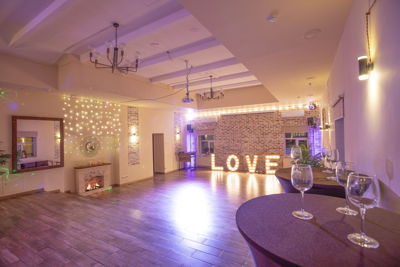 Арендовать Loft Lodge (Лофт Лодж) 7. Просторный уютный лофт. Просторное помещение с деревянным полом и светлыми стенами украшенными гирляндами. В углу электрический камин, напротив зеркало. На заднем плане кирпичная стена с крупными светящимися буквами «LOVE». Столы с бокалами для вина, потолочные светильники.