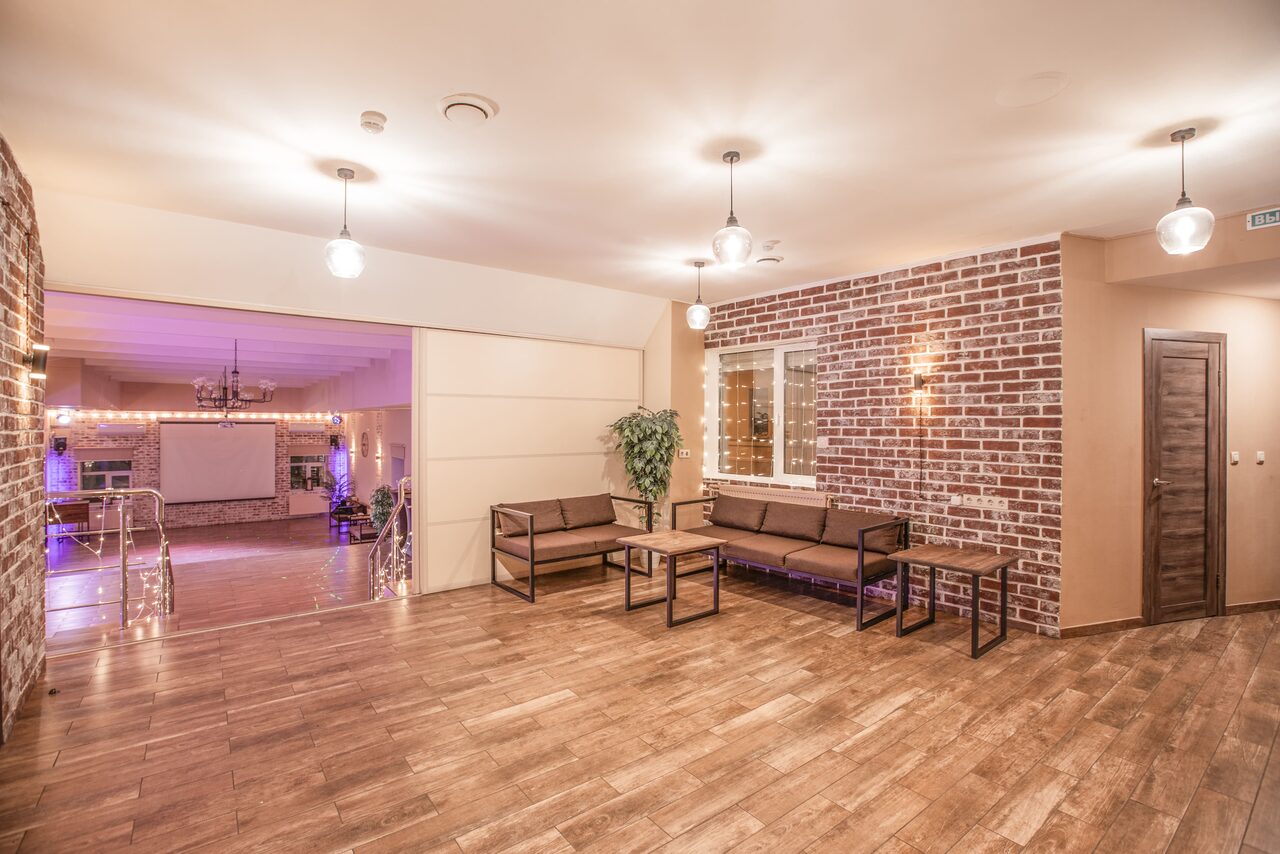 Арендовать Loft Lodge (Лофт Лодж) 7. Просторный уютный лофт. Просторное помещение с деревянным полом и кирпичными стенами. Угловой диван и кресла с журнальными столиками. Подвесные светильники и настенная лампа. Стена с окном, декоративное растение. Открытый проход в соседнее помещение с лестницей.