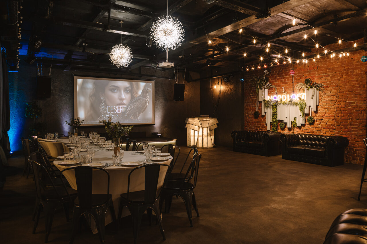 Арендовать Nord Loft(Норд Лофт). Пространство с кирпичной стеной, деревянным потолком и бетонным полом. В центре круглый стол с черными стульями, сервирован стеклянной посудой. На стене экран с изображением, рядом кожаные диваны и неоновая вывеска. Потолочные светильники и гирлянды создают мягкое освещение.