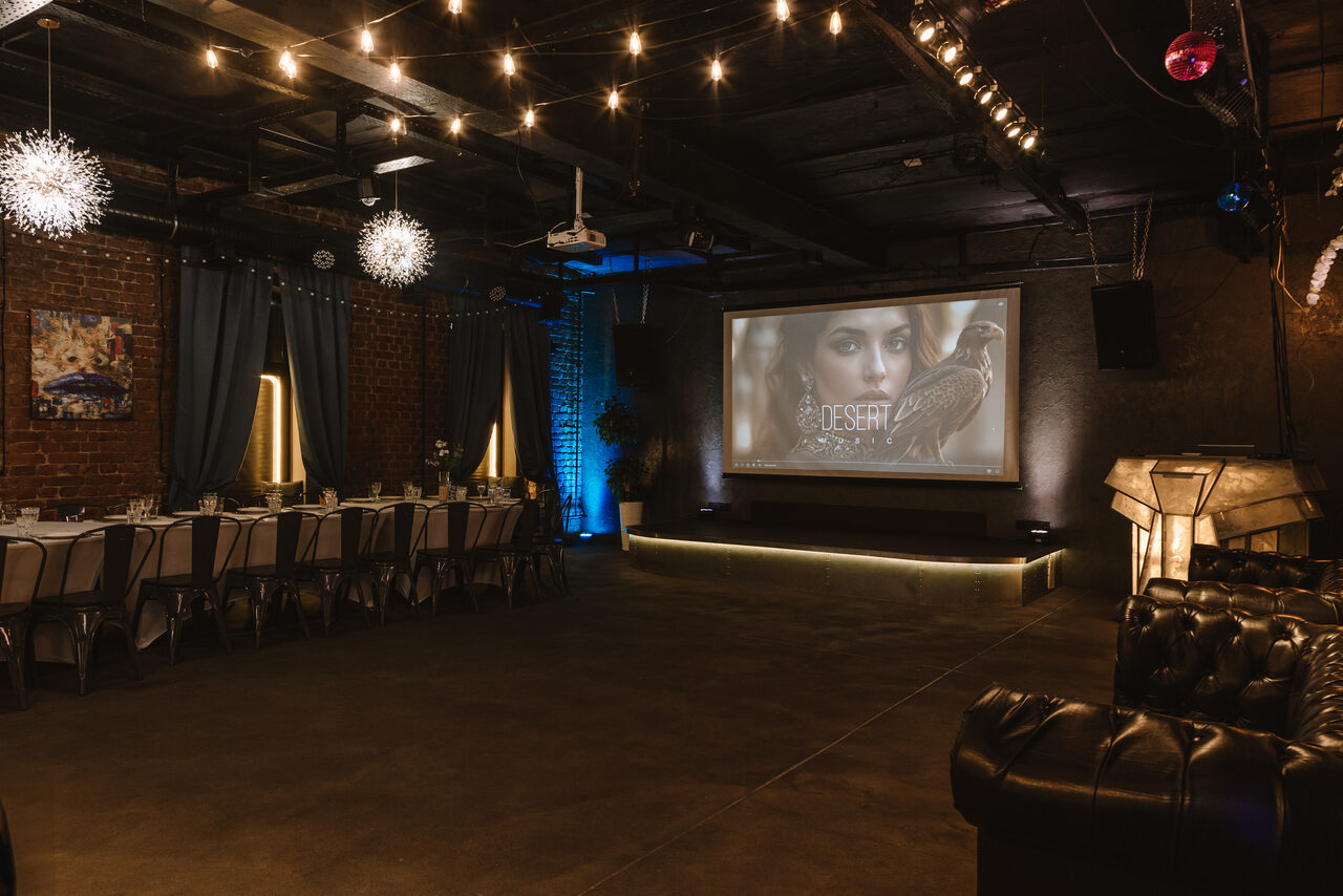 Арендовать Nord Loft(Норд Лофт). Просторное помещение с кирпичными стенами, длинный обеденный стол с металлическими стульями, кожаный диван, проектор и экран. Потолок украшен гирляндами, светильники-шары. Пол бетонный, освещение теплое, светодиодная подсветка синего и золотого оттенков.