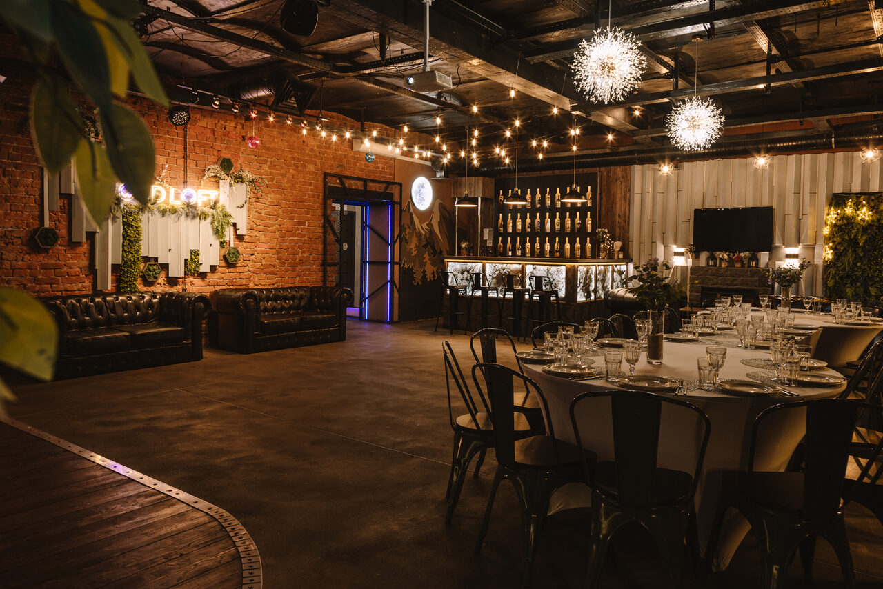 Арендовать Nord Loft(Норд Лофт). Большое помещение с кирпичными стенами, барной стойкой и обеденным столом. Кожаные диваны вдоль стены. Потолочные светильники и гирлянды создают мягкое освещение. Деревянный пол и металлические элементы интерьера.