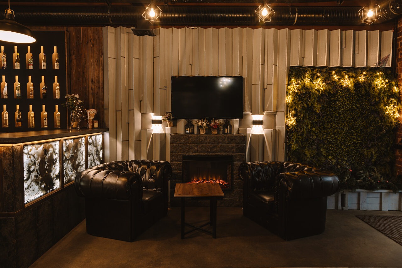 Арендовать Nord Loft(Норд Лофт). Лофт-пространство с кожаными креслами, деревянным столом, камином и телевизором на бежевой стене. Справа зелёная стена с подсветкой, слева барная стойка с бутылками и цветами, подвесные светильники, кирпич, дерево.