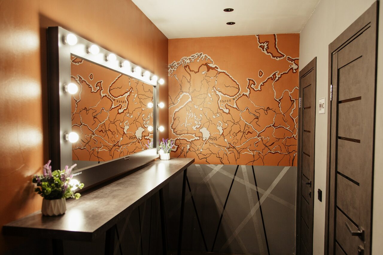 Арендовать Nord Loft(Норд Лофт). Гримерная с большой настенной картой; темная столешница с цветочным декором, большое зеркало в рамке с лампочками по периметру; стены коричневого и серого цветов; деревянные двери с горизонтальными вставками.