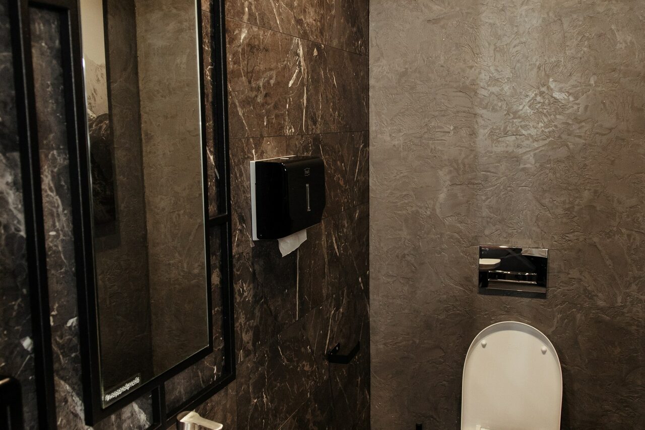 Арендовать Nord Loft(Норд Лофт). Туалетная комната с темными мраморными стенами, настенным унитазом, зеркалом в черной раме, черным диспенсером для туалетной бумаги и сенсорной панелью смыва.
