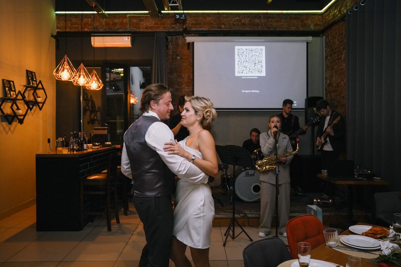 Арендовать Просторный лофт с видом на набережную. Пара танцует в зале с кирпичными стенами. Сцена слева, где играют музыканты. На заднем плане проектор с экраном. Справа обеденный стол с посудой. Светильники висят над барной стойкой. Шторы закрывают окна. Мягкое освещение, светло-коричневые и серые оттенки.