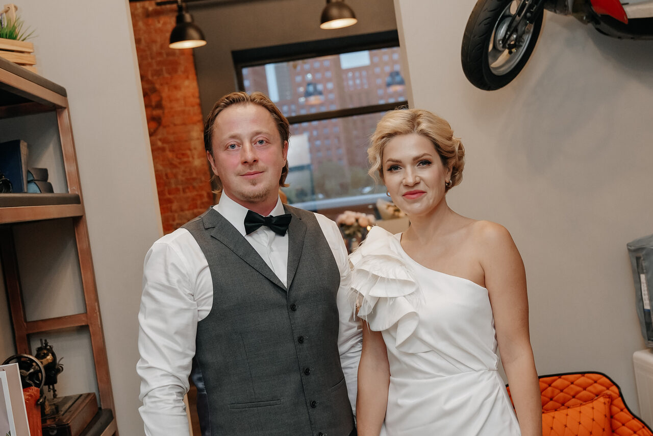 Арендовать Просторный лофт с видом на набережную. Лофт-пространство с кирпичной стеной, серыми и белыми поверхностями. Светильники с черными абажурами, окно с видом на городскую застройку. На стене подвешена мотоцикл, видна часть оранжевого кресла. На полке книги и декоративные элементы.