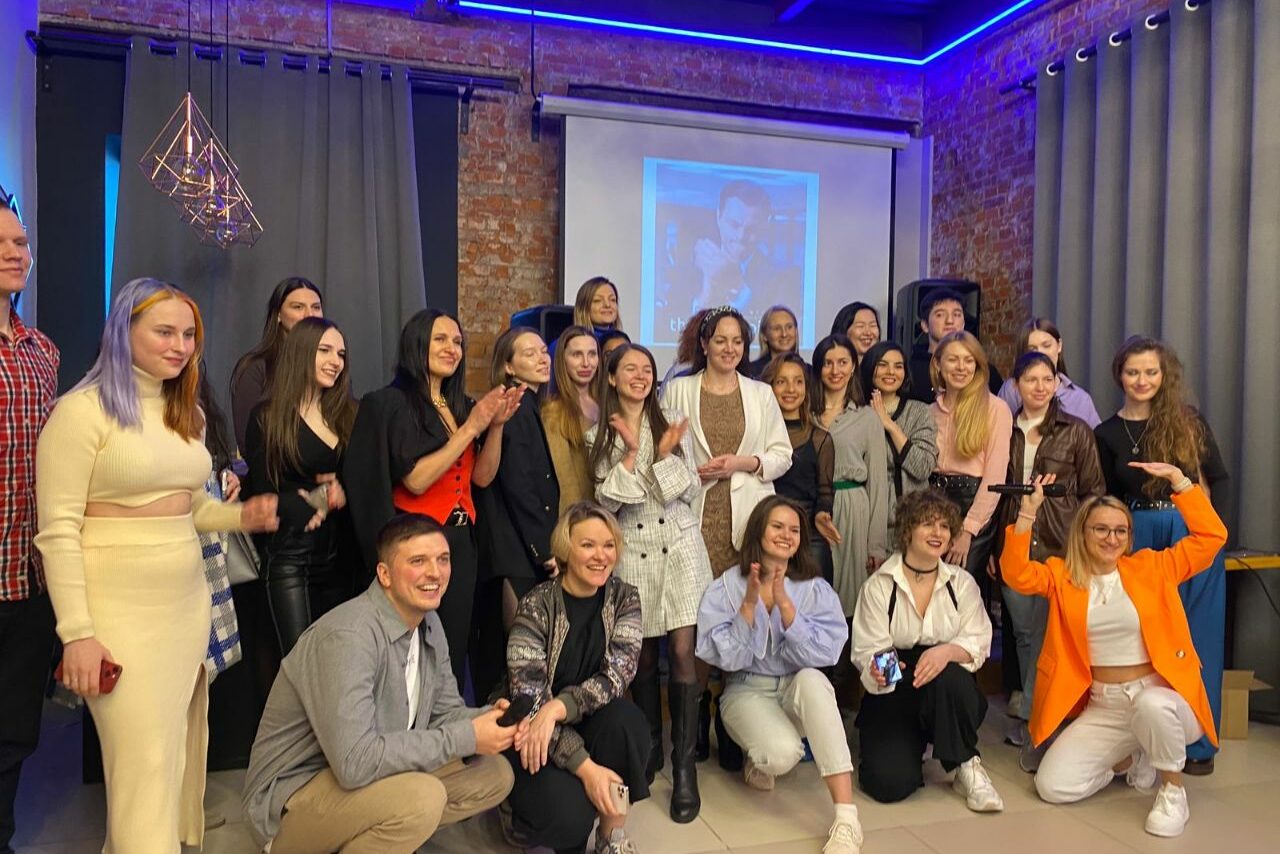 Арендовать Просторный лофт с видом на набережную. Группа людей в помещении с открытой кирпичной стеной, серыми шторами и проектором. Потолок с неоновыми световыми элементами. На полу светлая плитка. Рядом с проектором черные колонки. Люди одеты в разную повседневную и деловую одежду.