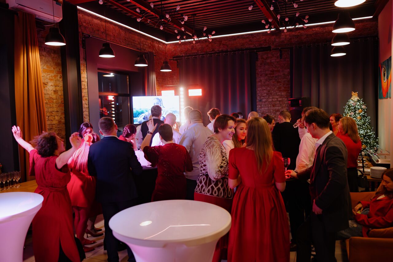 Арендовать Просторный лофт с видом на набережную. Лофт-пространство с кирпичными стенами и черным потолком. Выставлены круглые белые столы. Люди стоят в группе, некоторые в красной одежде. На стене рождественская елка. Освещение теплое, с подвесными лампами и светодиодной подсветкой. Завешенные окна.
