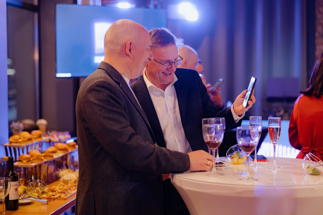 Арендовать Просторный лофт с видом на набережную. Стойка с закусками слева, круглый высокий стол с бокалами справа. Люди в официальной одежде, один держит смартфон, другой смотрит. Задник темный со световым экраном. В помещении тусклое освещение в оттенках синего и фиолетового.