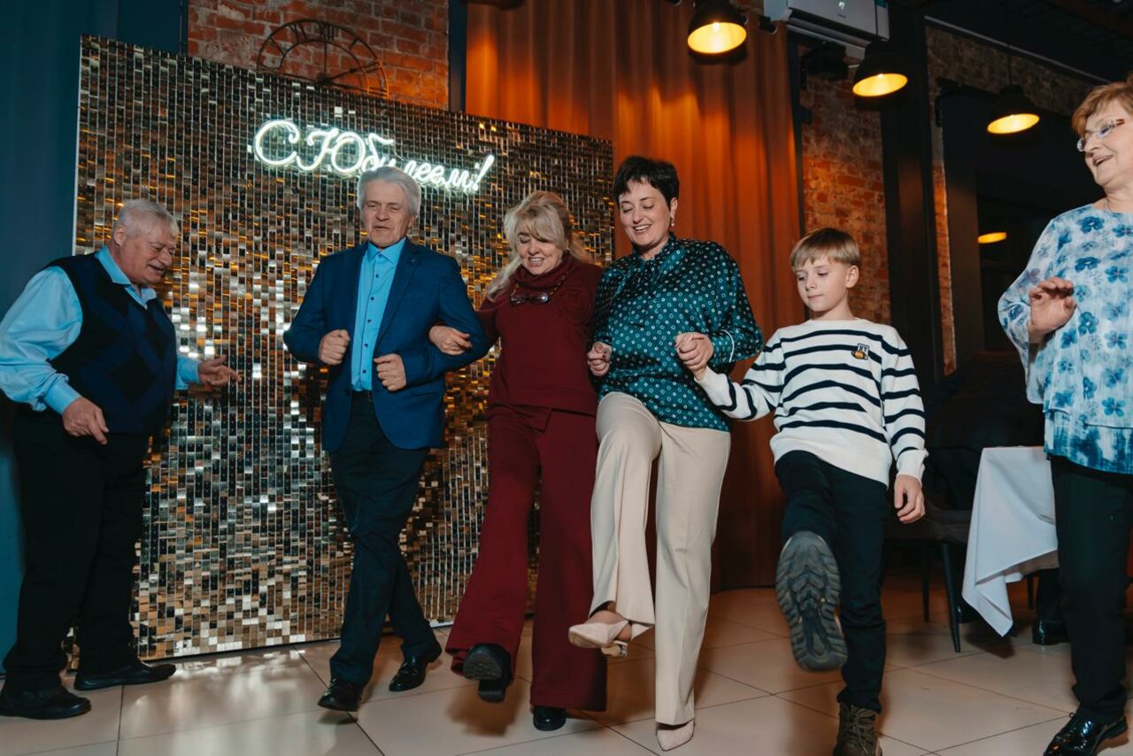 Арендовать Просторный лофт с видом на набережную. Группа людей танцует на светлом полу перед стеной с золотой зеркальной плиткой. На стене неоновая надпись. Справа видна часть накрытого стола. Фон из кирпичной стены и занавеса с софитами в теплых тонах.