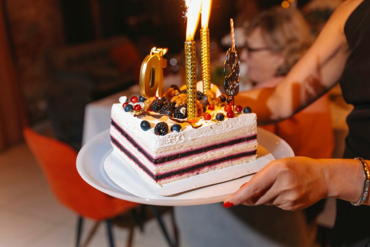 Арендовать Просторный лофт с видом на набережную. Торт на белой тарелке с ягодами и золотыми свечами в форме числа. На переднем плане руки человека, держащего тарелку. На заднем плане виден человек, сидящий за столом. Красный и бежевый цвета в обстановке.