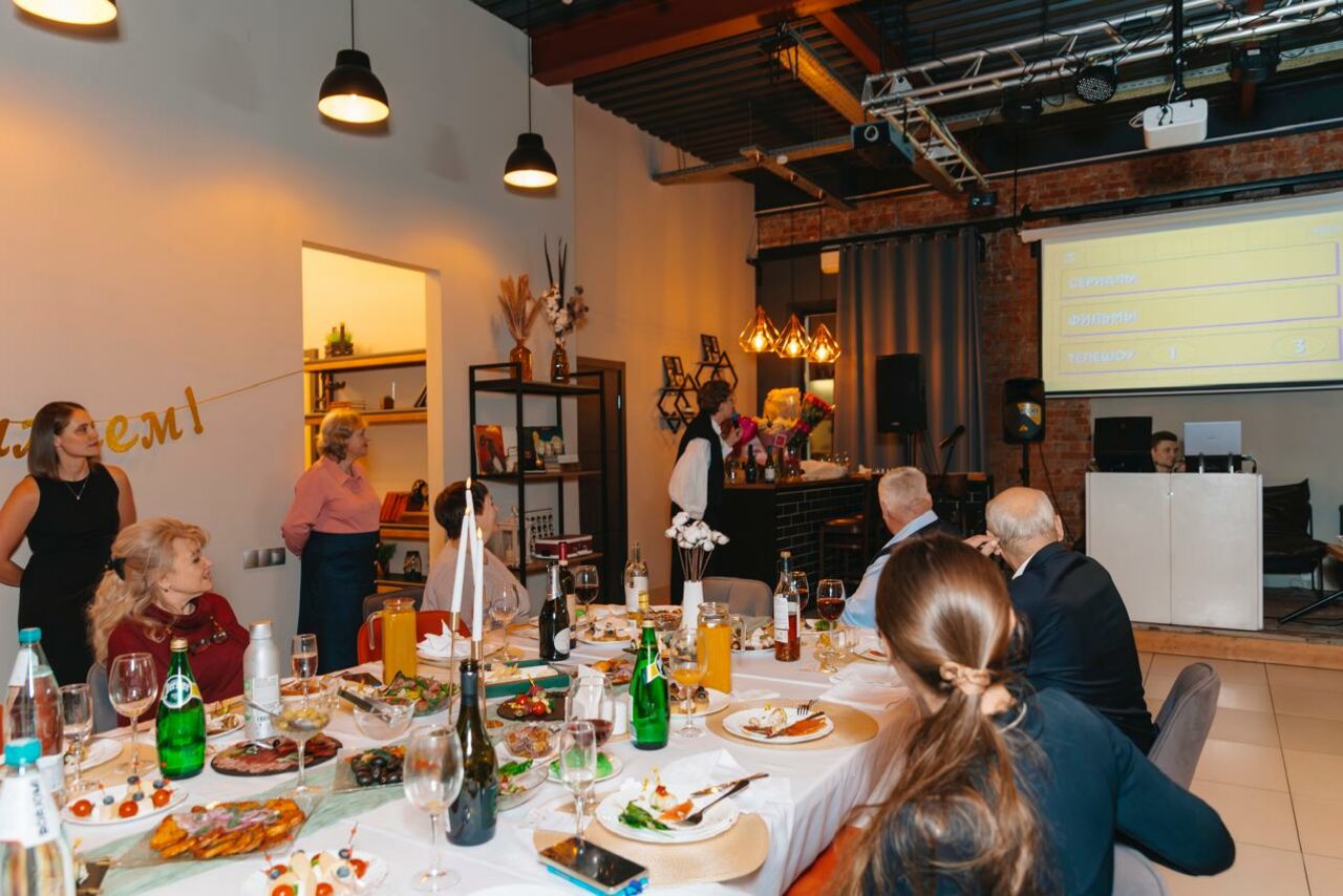 Арендовать Просторный лофт с видом на набережную. Просторное помещение с кирпичной стеной, подвесные светильники, столы с белыми скатертями и накрытыми блюдами, стулья. Оборудование: проектор с экраном, звуковая аппаратура. На полках книги и декор. Люди в формальной одежде. Надпись на стене: "welcсome".