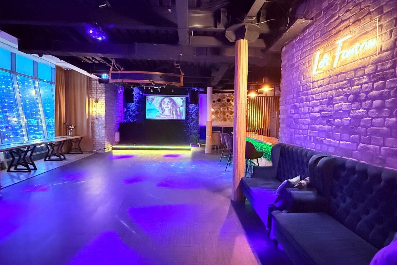 Арендовать Лофт Фантом. Пространство в стиле лофт с кирпичными стенами, неоновыми светильниками и деревянными элементами. На стене проектор, рядом с диваном стулья. Быстрые столы вдоль окна, занавешенные шторами. Потолочные светильники создают разноцветное освещение.