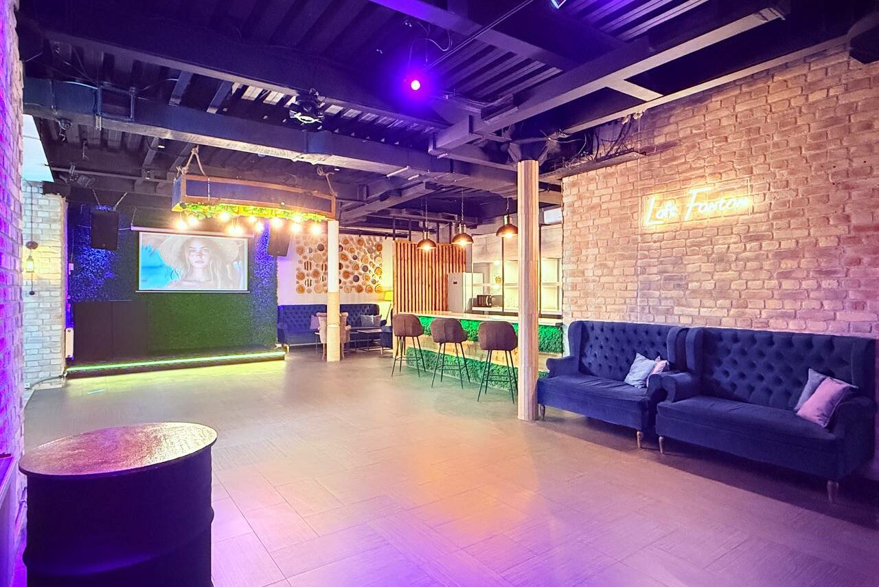 Арендовать Лофт Фантом. Просторное помещение с открытой планировкой, кирпичные стены, барная стойка с высокими стульями, синий диван, неоновая вывеска, большой экран для проекций. Потолок темный с открытыми балками, комбинированное освещение. Пол плиточный.