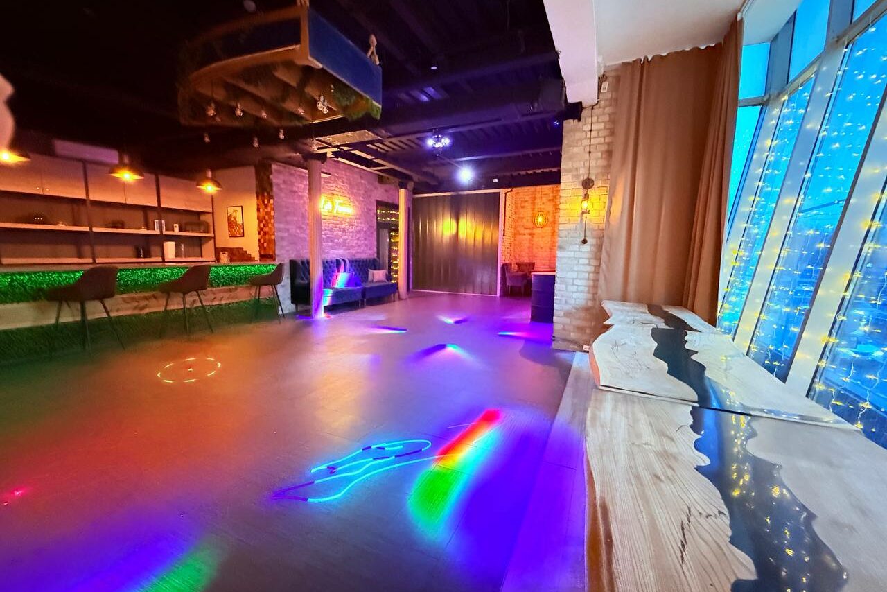 Арендовать Лофт Фантом. Просторное помещение с барной стойкой, высокими табуретами и открытыми полками. На полу разноцветные световые эффекты. Окна украшены светодиодными гирляндами. Стены из кирпича, потолок темный, длинный деревянный стол у окна.