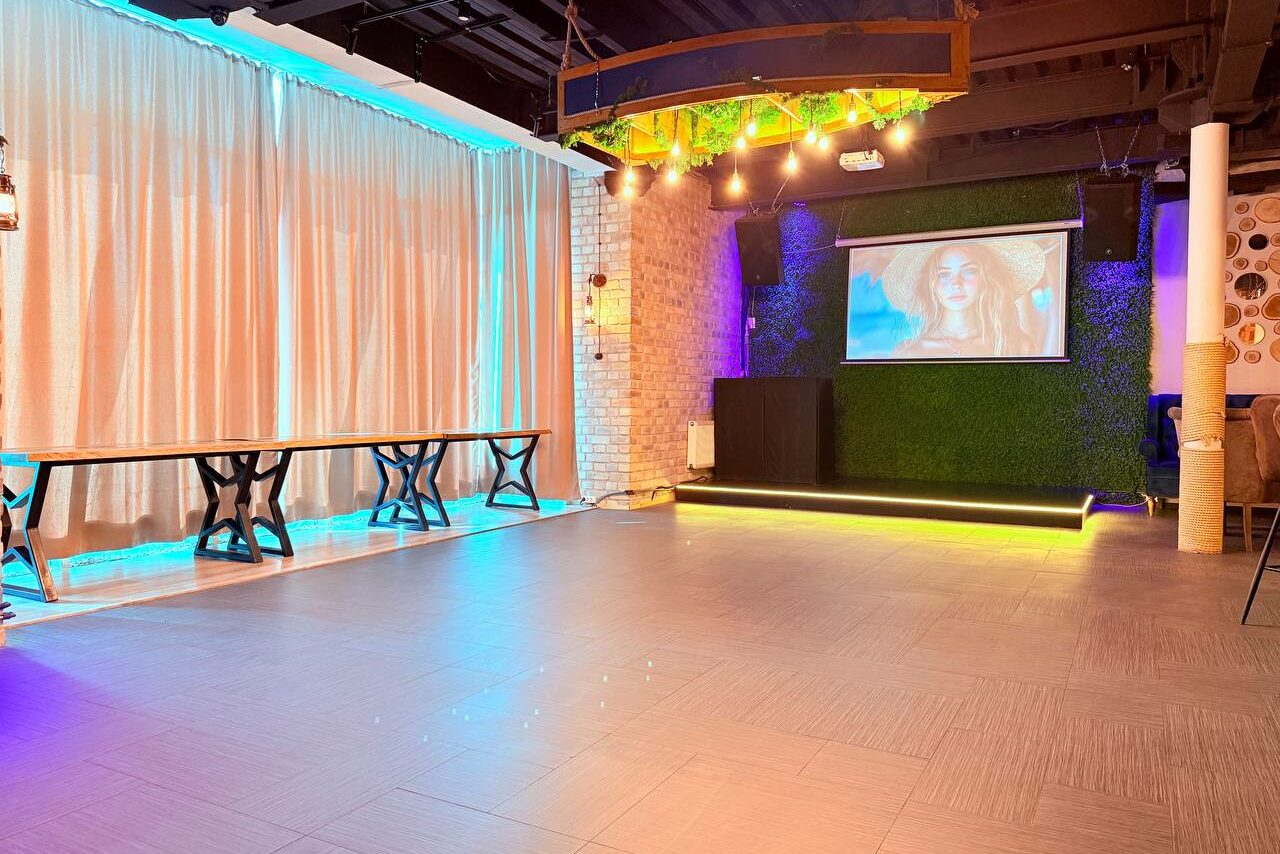 Арендовать Лофт Фантом. Просторное помещение с занавесками, кирпичными стенами и светлым плиточным полом. Включает три деревянных стола с металлическими ножками, проектор на стене с зеленым покрытием, яркое разноцветное освещение и декоративные элементы на потолке.