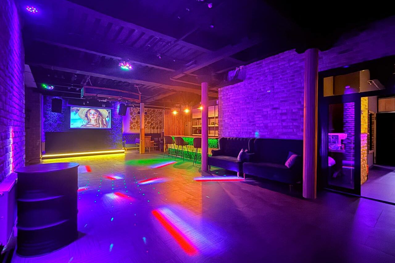Арендовать Лофт Фантом. Лофт-пространство с кирпичными стенами, сценой и экраном. Черные диваны вдоль стены, стойка с зелеными барными стульями, дискотечное освещение с яркими цветами. Потолок с открытыми балками, деревянные элементы. Табличек или знаков не видно.