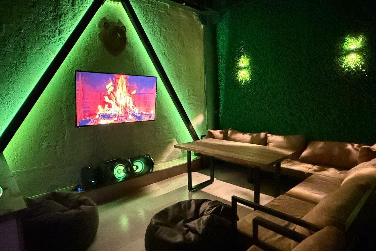 Арендовать Лофт Фантом. Лофт-пространство с диваном и креслами, деревянный стол. Телевизор на стене показывает камин. На полу пуфы. Зеленая подсветка и стена с растительностью. Потолок с белыми балками. Стена сделана из штукатурки.