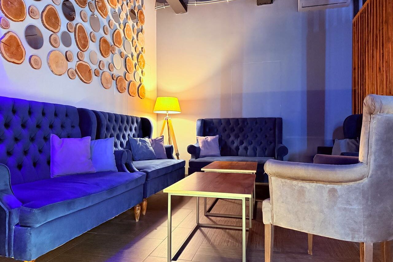 Арендовать Лофт Фантом. Лофт-пространство с мягкими синими диванами и креслом, деревянным столом, лампой с абажуром. Стена украшена круглым деревянным декором. Пол темный, освещение теплое.