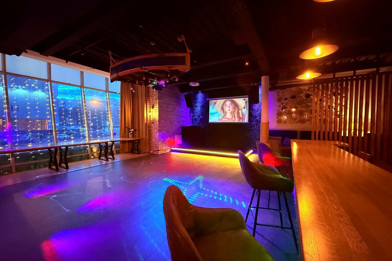 Арендовать Лофт Фантом. Просторное помещение с панорамными окнами и тематическим освещением. Длинный деревянный стол с высокими разноцветными стульями, барная стойка, сцена с экраном, кирпичная и деревянная отделка. Потолочные светильники. Подвесные декорации.