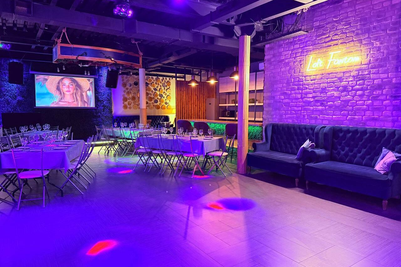 Арендовать Лофт Фантом. Пространство с синим неоновым освещением, кирпичными стенами, банкетными столами и стульями. Экран с изображением, диваны темного цвета, барная стойка с подсветкой. На стене неоновая надпись. Потолок темный с открытыми коммуникациями.