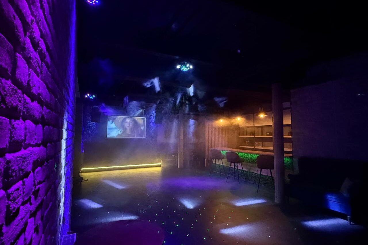 Арендовать Лофт Фантом. Тёмное помещение с кирпичной стеной, проектор с изображением, неоновая подсветка, барная стойка с высокими стульями, цветные световые эффекты на полу, дымка в воздухе.