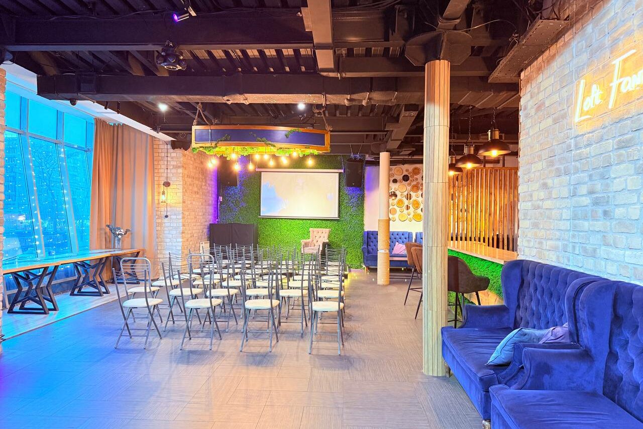 Арендовать Лофт Фантом. Просторное помещение с кирпичными стенами, большими окнами и веревочными шторами. Ряд стульев перед экраном и проекционным оборудованием. Декор из зелени, подиум, яркое освещение и неоновые вывески. Синие диваны, деревянные и металлические элементы.