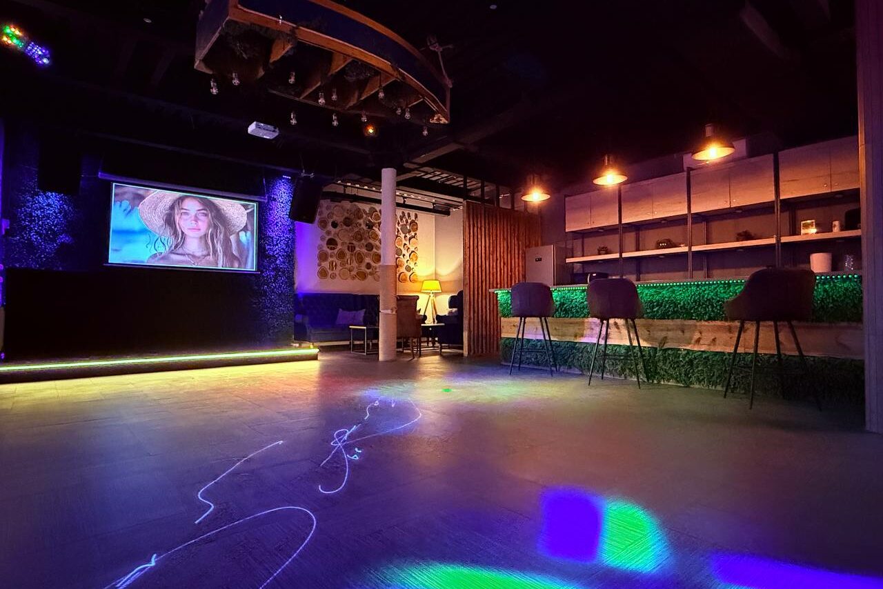 Арендовать Лофт Фантом. Пространство с проектором на стене, барная стойка с высокими стульями, неоновая подсветка, черный потолок с прожекторами, одинокий торшер, светлые деревянные полы, зелёные элементы декора, тёмные полки, светодиодное освещение на полу.