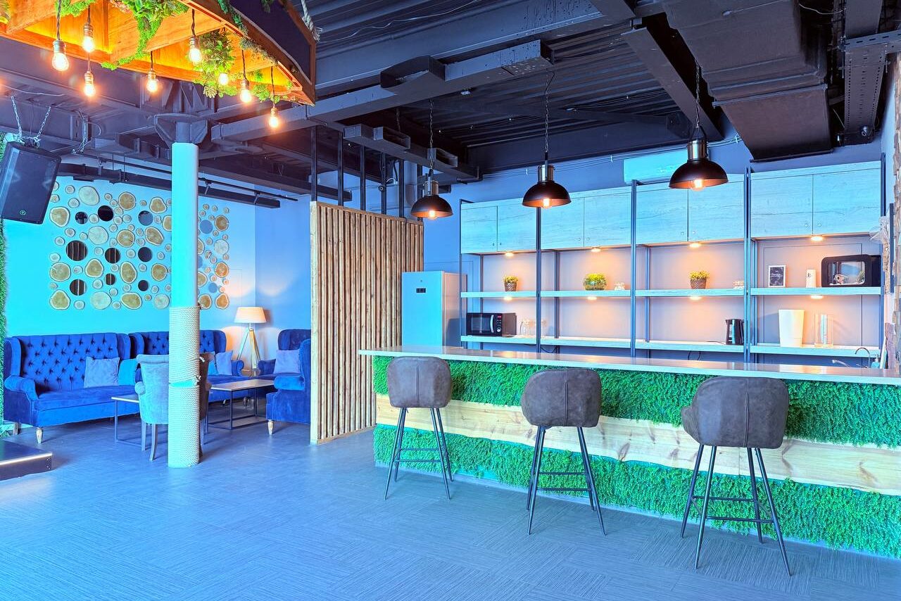 Арендовать Лофт Фантом. Современное пространство с барной стойкой, декором из искусственной травы, тремя барными стульями. Справа стеллажи с подсветкой, слева зона отдыха с синими диванами и декором на стене. Потолок с открытыми коммуникациями и подвесными светильниками.