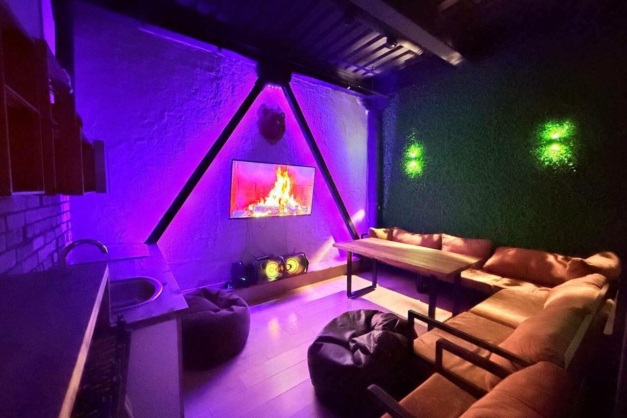 Арендовать Лофт Фантом. Лофт-пространство с диваном и креслами, деревянные полы, телевизор с изображением камина на стене, неоновая подсветка фиолетового и зеленого цветов, стена с искусственными растениями, барная стойка с раковиной.