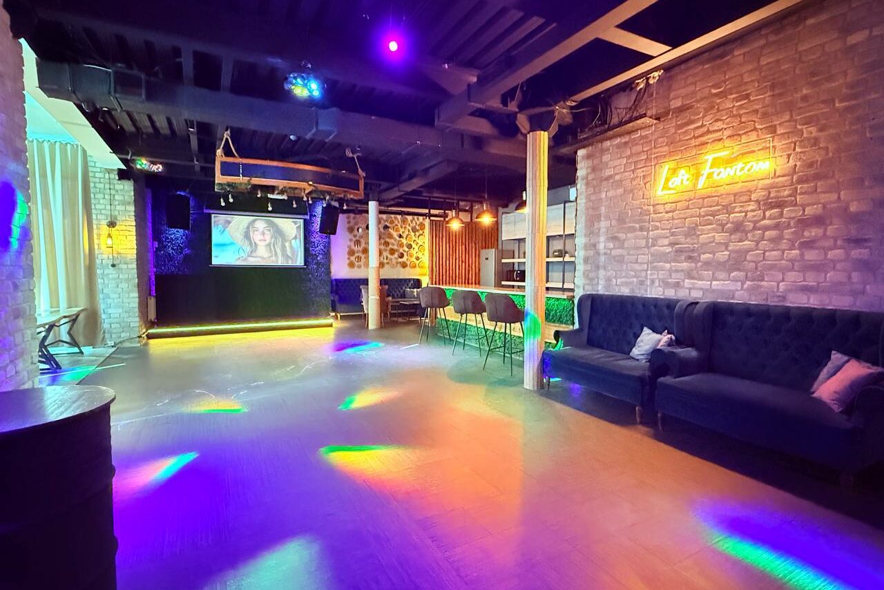 Арендовать Лофт Фантом. Просторное помещение с кирпичными стенами и деревянными балками. На полу паркет, разноцветное освещение. Слева зона с креслами и столами. Справа темно-синий диван. На стене светящаяся вывеска. На задней стене экран и колонки.