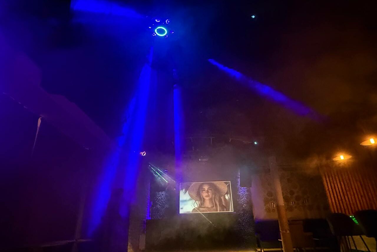 Арендовать Лофт Фантом. Помещение с цветным освещением, в центре экран с изображением. Световые лучи, искусственный дым. Тёмная обстановка, на стенах декоративные элементы. Освещение из синих прожекторов и нескольких тёплых ламп.