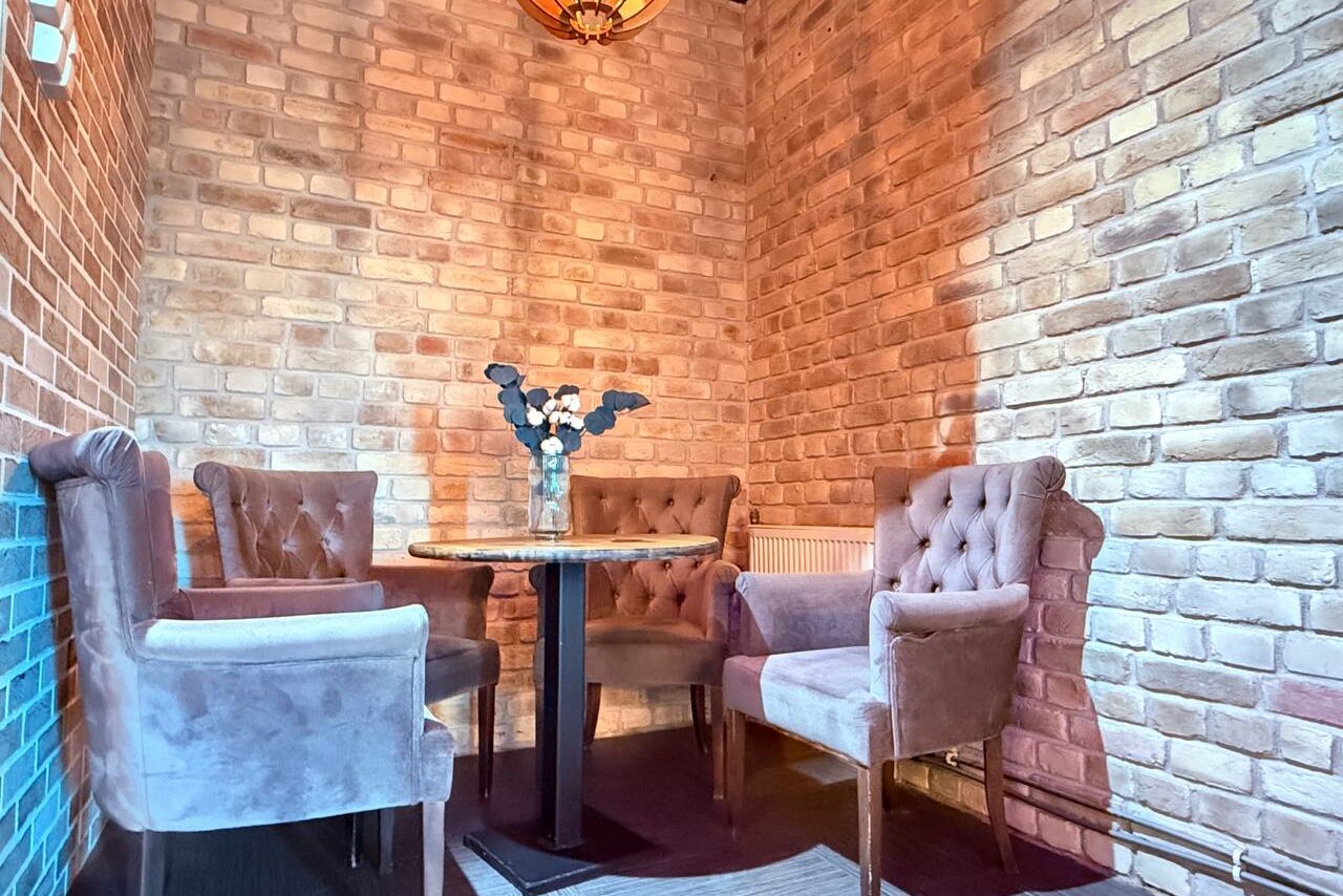 Арендовать Лофт Фантом. Небольшая комната с кирпичными стенами, круглым столом и четырьмя мягкими креслами бежевого цвета. На столе прозрачная ваза с сухими цветами. Освещение теплое, присутствует подвесной светильник. Пол темный, видна часть ковра.