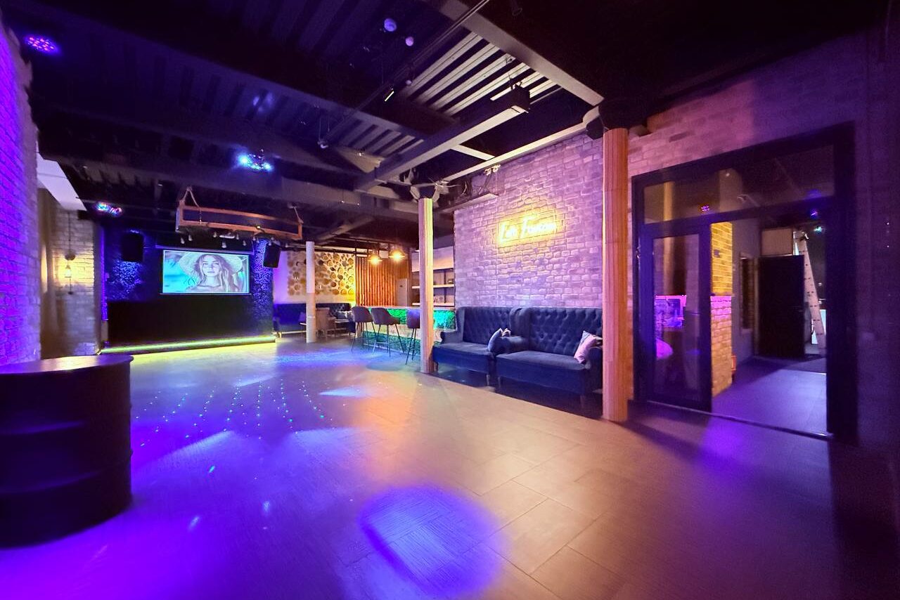Арендовать Лофт Фантом. Лофт-пространство с кирпичными стенами и черным потолком. Большой экран, сцена. В центре столы и стулья, вдоль стены синий диван. Неоновая вывеска. Пестрое освещение с фиолетовыми и желтыми акцентами. Пол темный, плиточный.