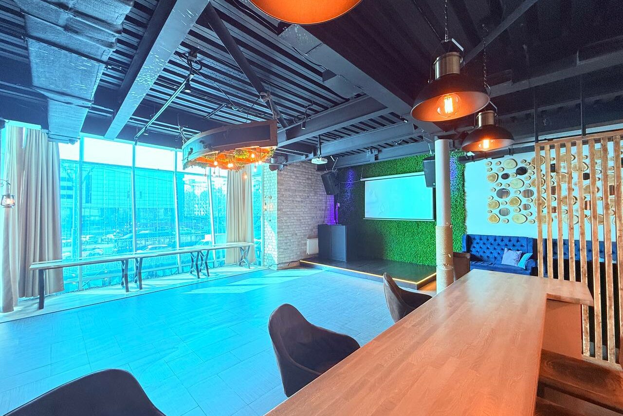 Арендовать Лофт Фантом. Просторное помещение с большими окнами, столом и стульями, зоной отдыха с диваном, декором из зелени и кирпичной стены. Светильники в индустриальном стиле, дерево и металл в оформлении, нейтральные и теплые тона в интерьере.