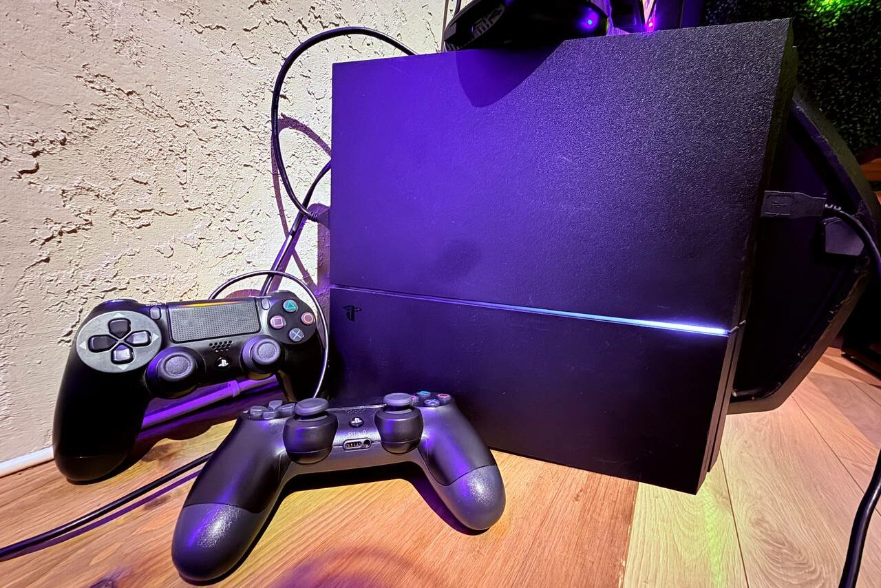 Арендовать Лофт Фантом. Игровая консоль и два геймпада черного цвета на деревянном полу у текстурированной стены.