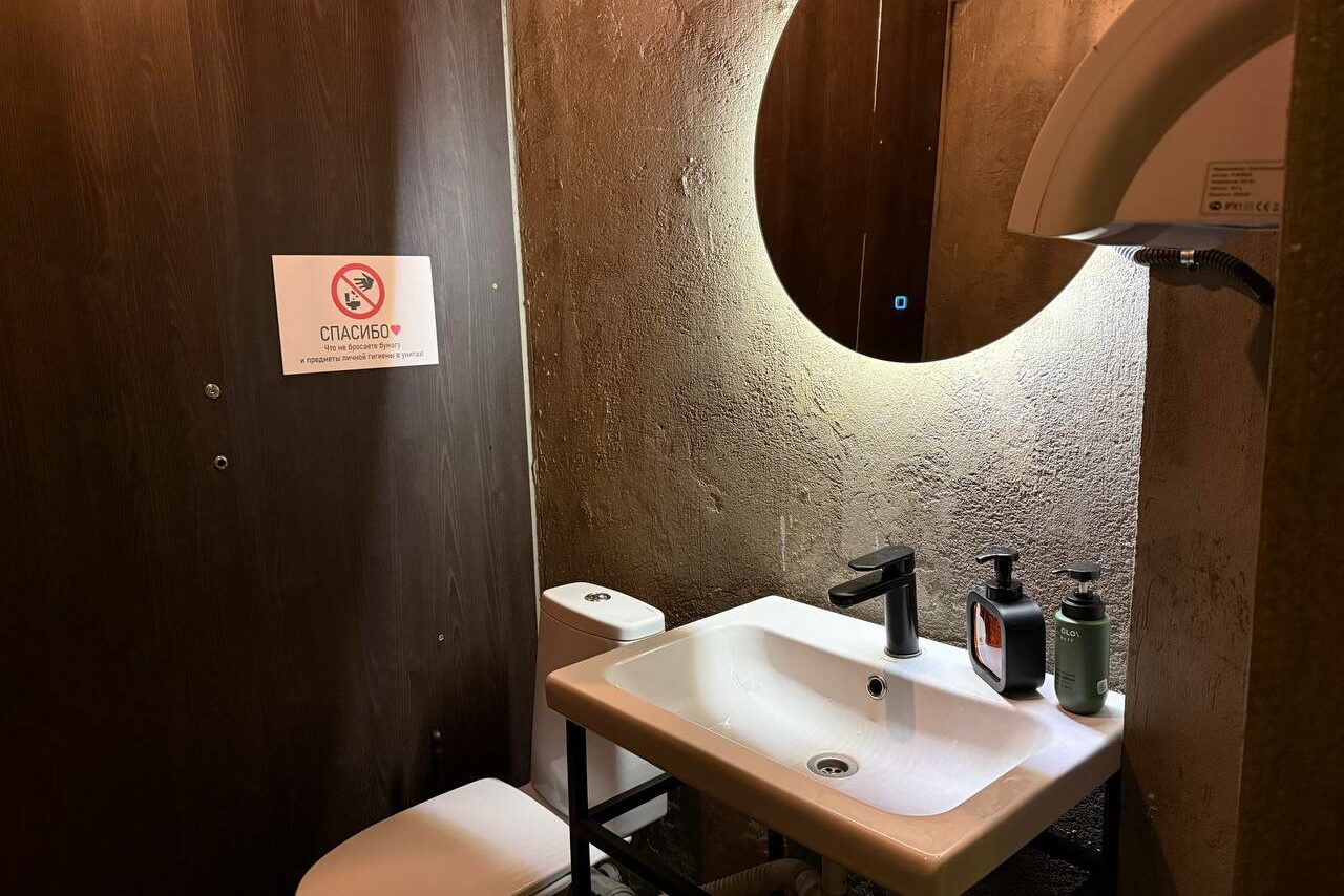 Арендовать Лофт Фантом. Туалетное помещение с серыми бетонными стенами. Угловая раковина с черным смесителем и рядом жидкое мыло. Над раковиной круглое зеркало с подсветкой. Слева на деревянной стене знак с текстом. Белый унитаз, сушилка для рук.