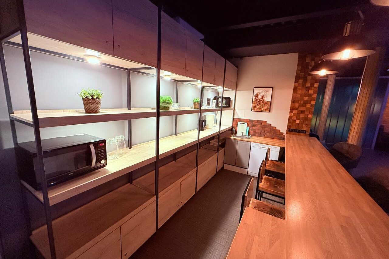 Арендовать Лофт Фантом. Кухонное пространство с длинной деревянной барной стойкой и табуретами, полки с освещением и растениями, микроволновая печь, стеклянные и керамические емкости, картина на стене, светлые деревянные и металлические конструкции, темный пол, кирпичная кладка.