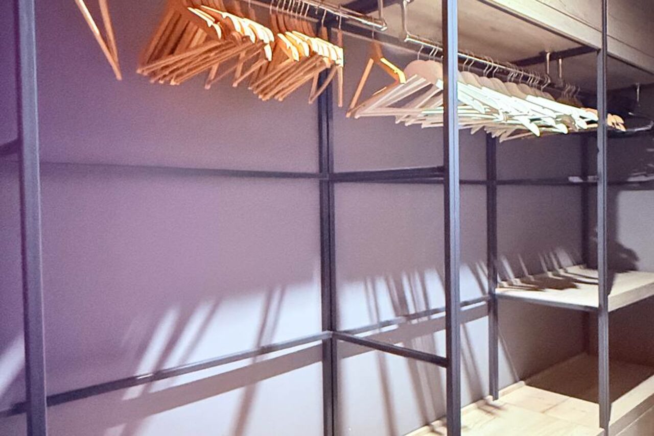 Арендовать Лофт Фантом. Гардеробная комната с металлическими стойками и множеством деревянных и пластиковых вешалок. Стены и пол светлых тонов. Яркое освещение.