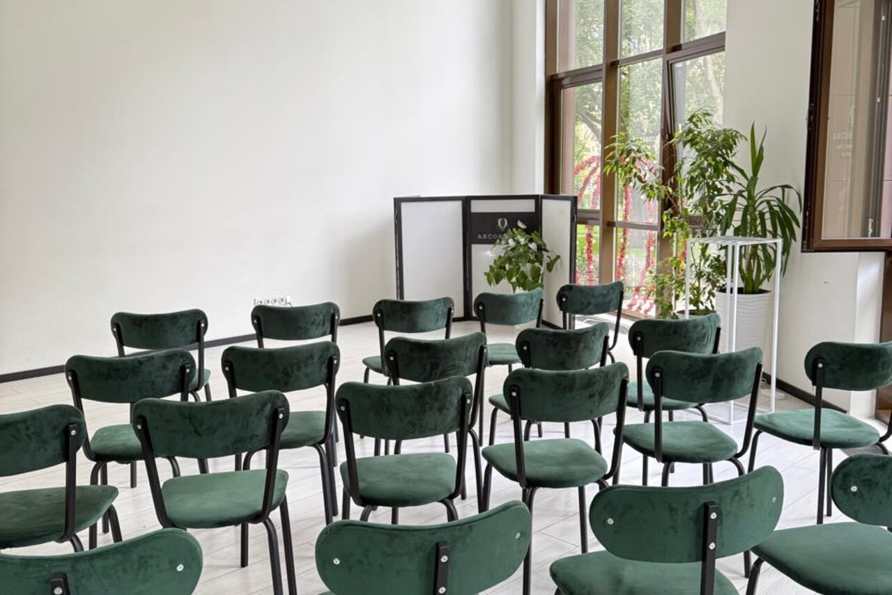 Арендовать Arcobaleno Loft(Аркобалено Лофт). Просторное помещение с белыми стенами и большими окнами. Ряд зеленых стульев с черными каркасами, напротив небольшой трибуны. На подоконнике стоят растения в горшках. Пол светлый.