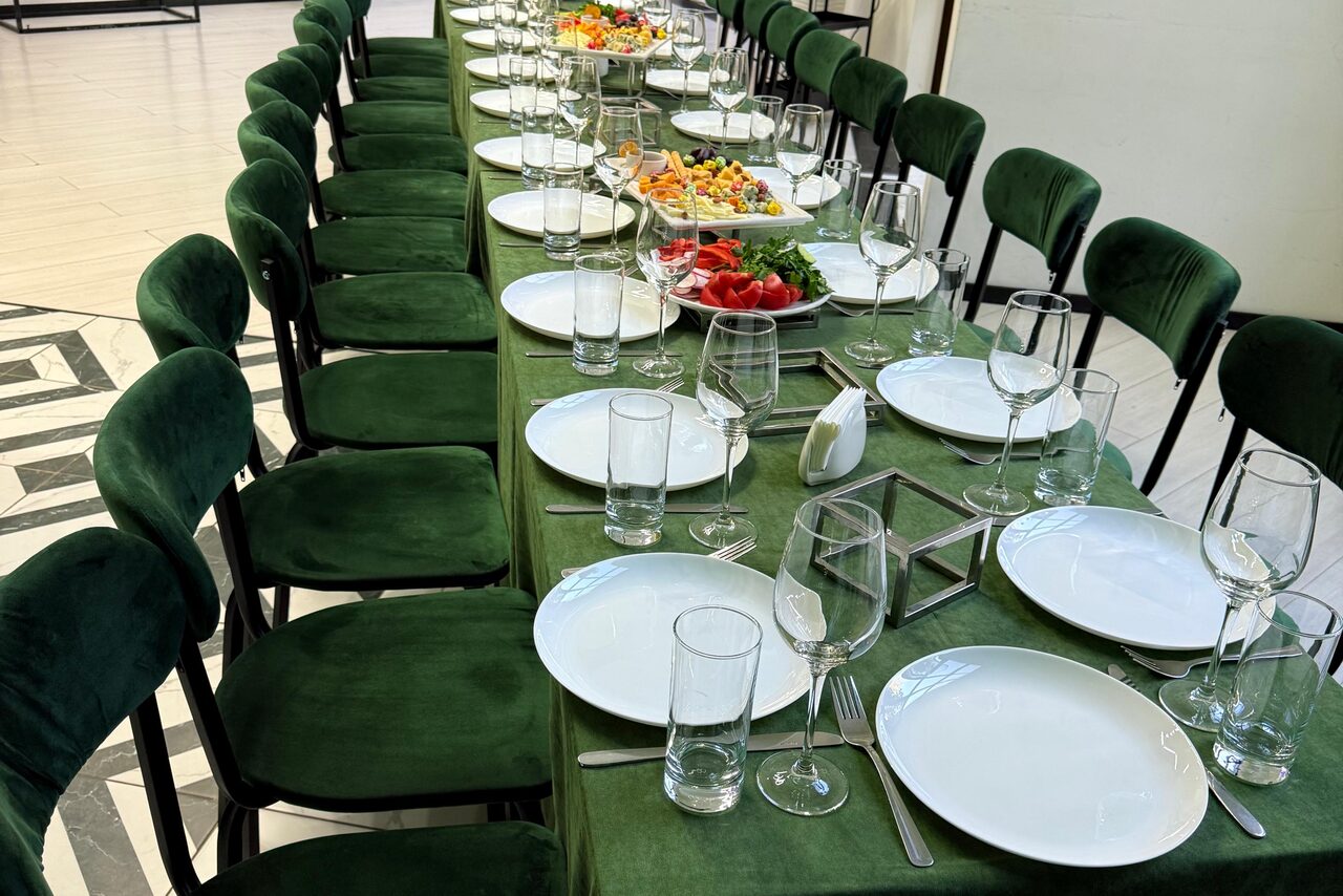 Арендовать Arcobaleno Loft(Аркобалено Лофт). Длинный стол, накрытый скатертью зелёного цвета, окружён стульями с зелёной обивкой. На столе расставлены белые тарелки, бокалы и стаканы, приборы. Разложены салаты и овощи. Пол выложен плиткой с черно-белым рисунком. Освещение естественное.