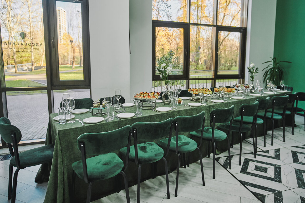 Арендовать Arcobaleno Loft(Аркобалено Лофт). Просторное помещение для мероприятий с длинным столом, покрытым зелёной скатертью, и зелёными стульями. На столе посуда и закуски. Широкие окна с видом на деревья. Черно-белый узор на полу. Светлые стены.