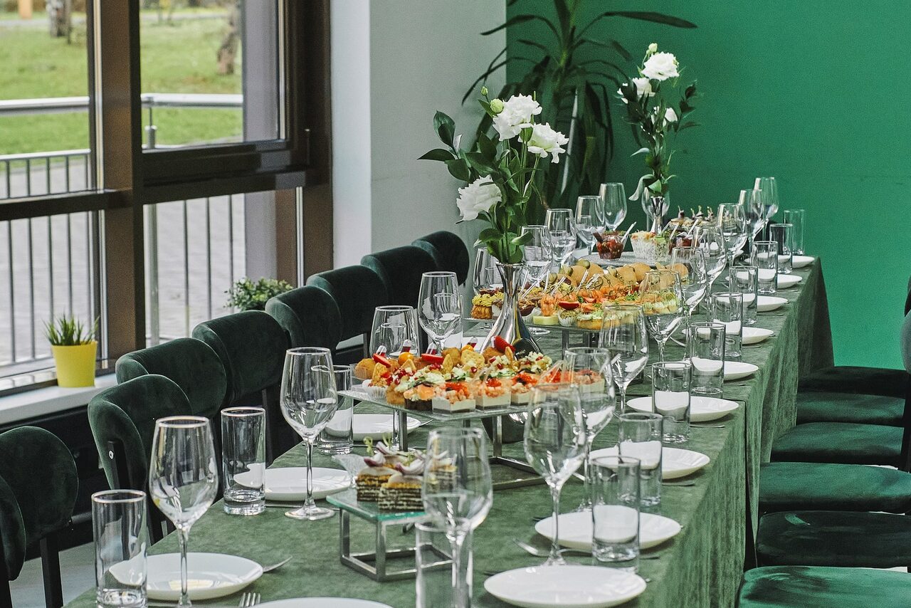 Арендовать Arcobaleno Loft(Аркобалено Лофт). Пространство для мероприятий с длинным столом, покрытым зелёной скатертью. На столе расставлены бокалы, тарелки и закуски. Стулья обиты зелёной тканью. У стен декоративные растения и крупные окна, через которые видно улицу.