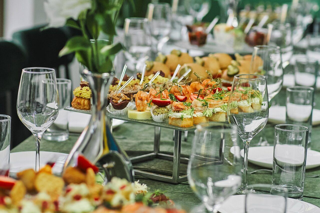 Арендовать Arcobaleno Loft(Аркобалено Лофт). Длинный обеденный стол, покрытый зелёной скатертью, с расставленными белыми тарелками и стеклянной посудой. В центре стола на подставке расположено разнообразное канапе. На заднем плане видна зелень в вазе.