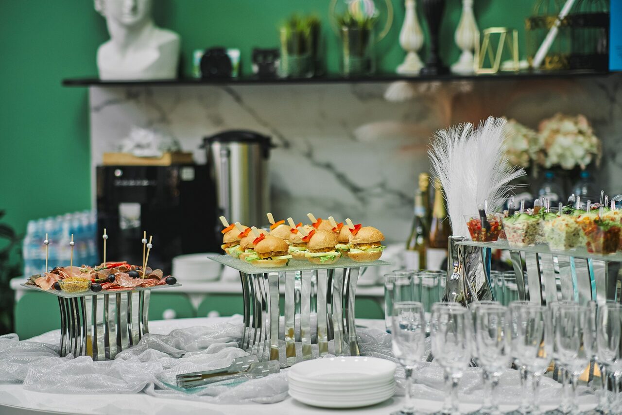 Арендовать Arcobaleno Loft(Аркобалено Лофт). Стол с закусками на подставках, ряды бокалов и белые тарелки. Задний план: кофемашина, бутылки, мраморная стена, декор, зелёные стены, вазы и статуэтка.