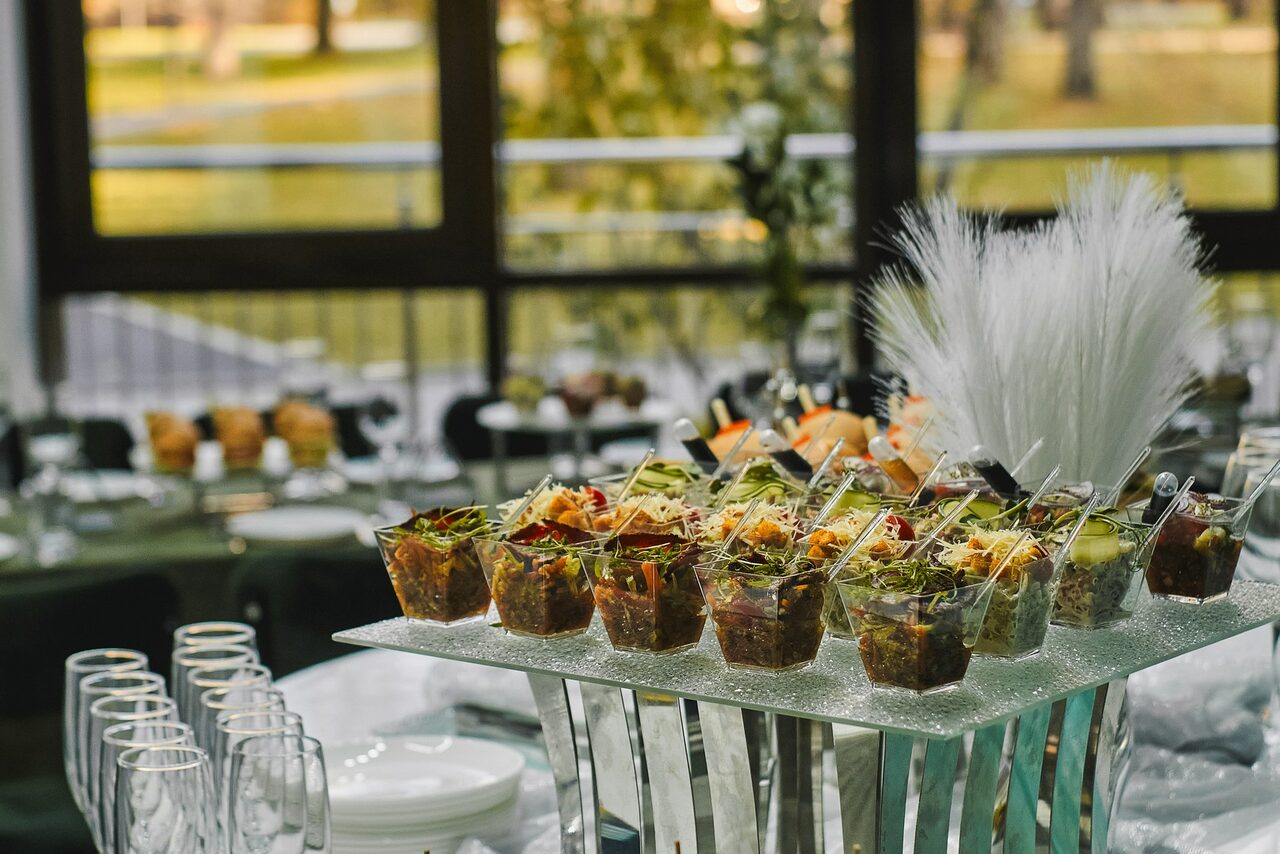 Арендовать Arcobaleno Loft(Аркобалено Лофт). Стол в помещении, оформлен для мероприятия. На переднем плане подносы с закусками в стаканах. На столе стеклянные бокалы и пустые тарелки. Панорамные окна пропускают естественный свет. На заднем плане видны элементы растения и декора.