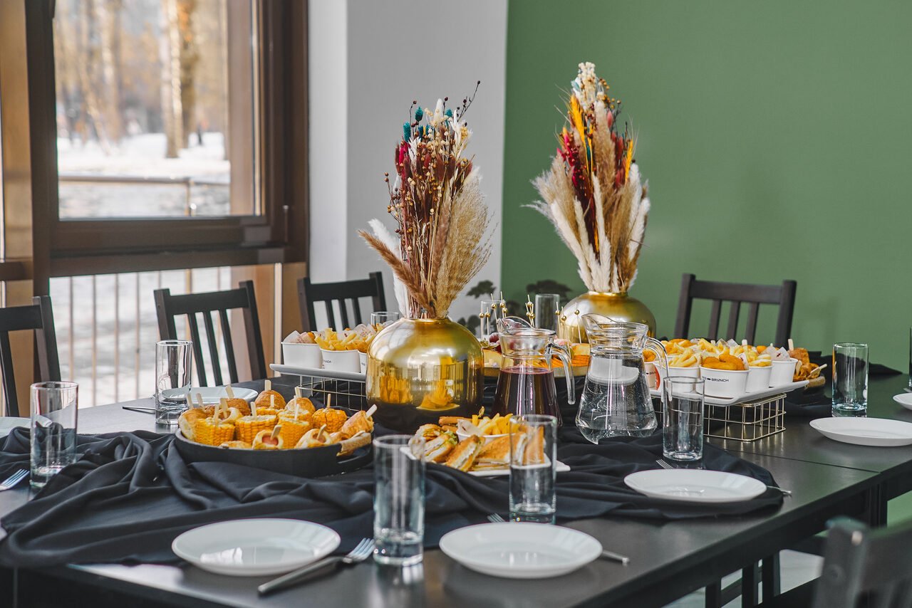 Арендовать Arcobaleno Loft(Аркобалено Лофт). Столовая зона с черными стульями и столом, сервированным белыми тарелками и стеклянными стаканами. По центру композиции из сухоцветов в золотых вазах, рядом закуски в корзинах. Задняя стена зеленого цвета. Панорамное окно.