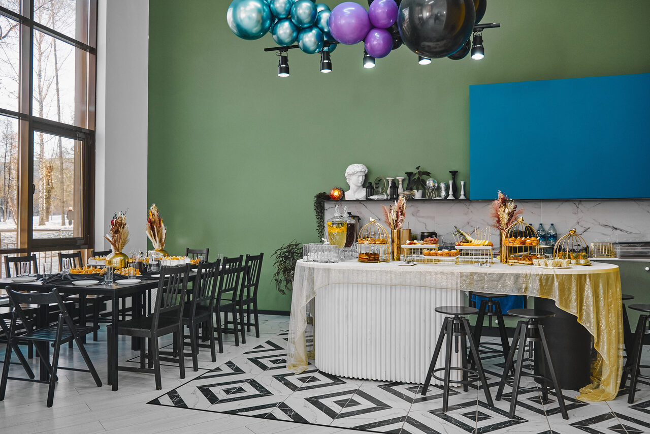 Арендовать Arcobaleno Loft(Аркобалено Лофт). Просторное помещение с сервированным столом для фуршета. На стене полка с декоративными элементами. Стены зелёного и синего цвета, пол белый с узором. Подвесные разноцветные шары. чёрные стулья, большие окна, естественный свет.